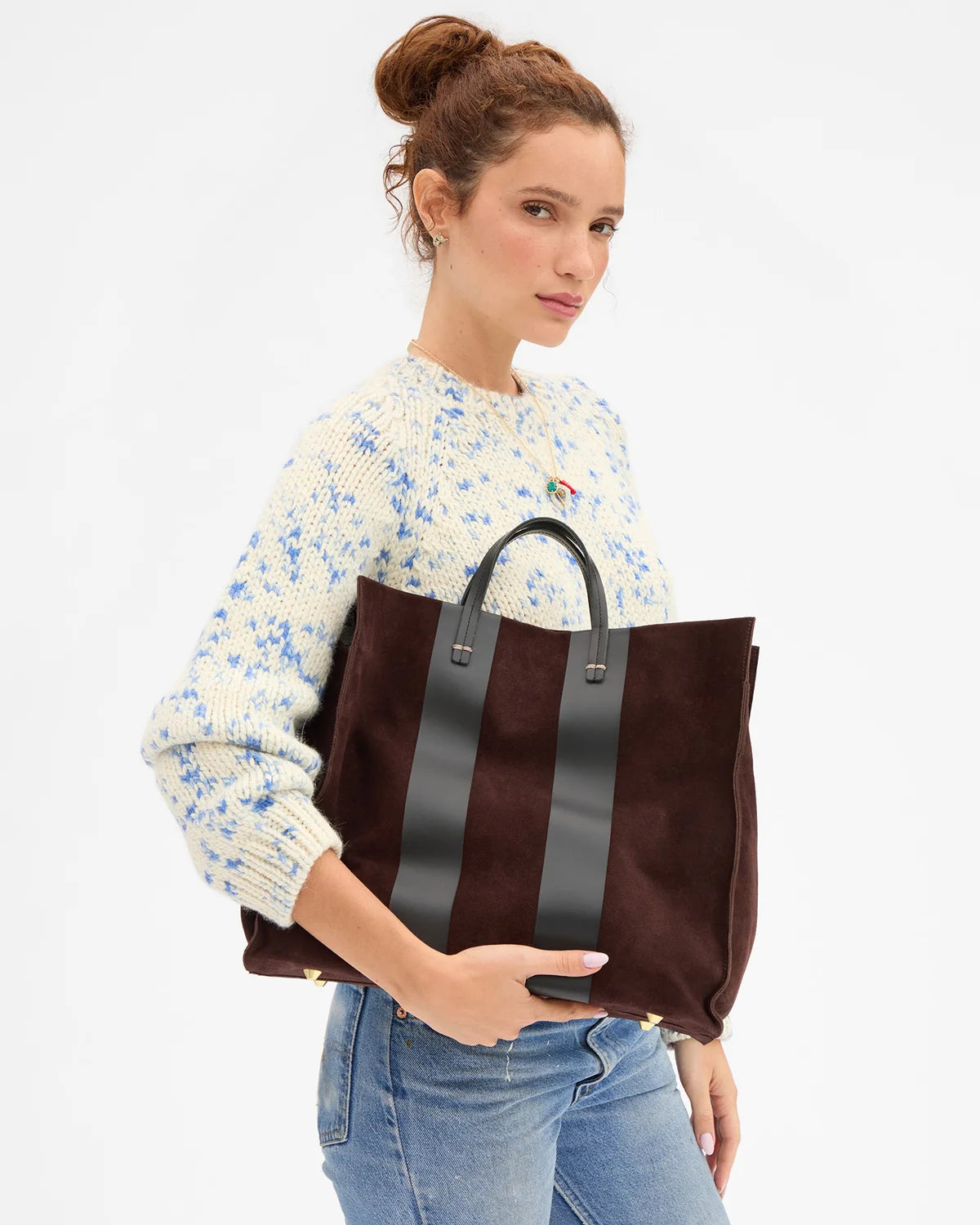 A woman wearing a cream knit sweater with blue floral patterns and blue jeans holds a large dark brown suede tote bag with black leather handles and vertical black leather stripes against a plain white background.