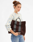 A woman wearing a cream knit sweater with blue floral patterns and blue jeans holds a large dark brown suede tote bag with black leather handles and vertical black leather stripes against a plain white background.