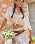 Woman in a white lace outfit standing outdoors with a stone wall and plants in the background