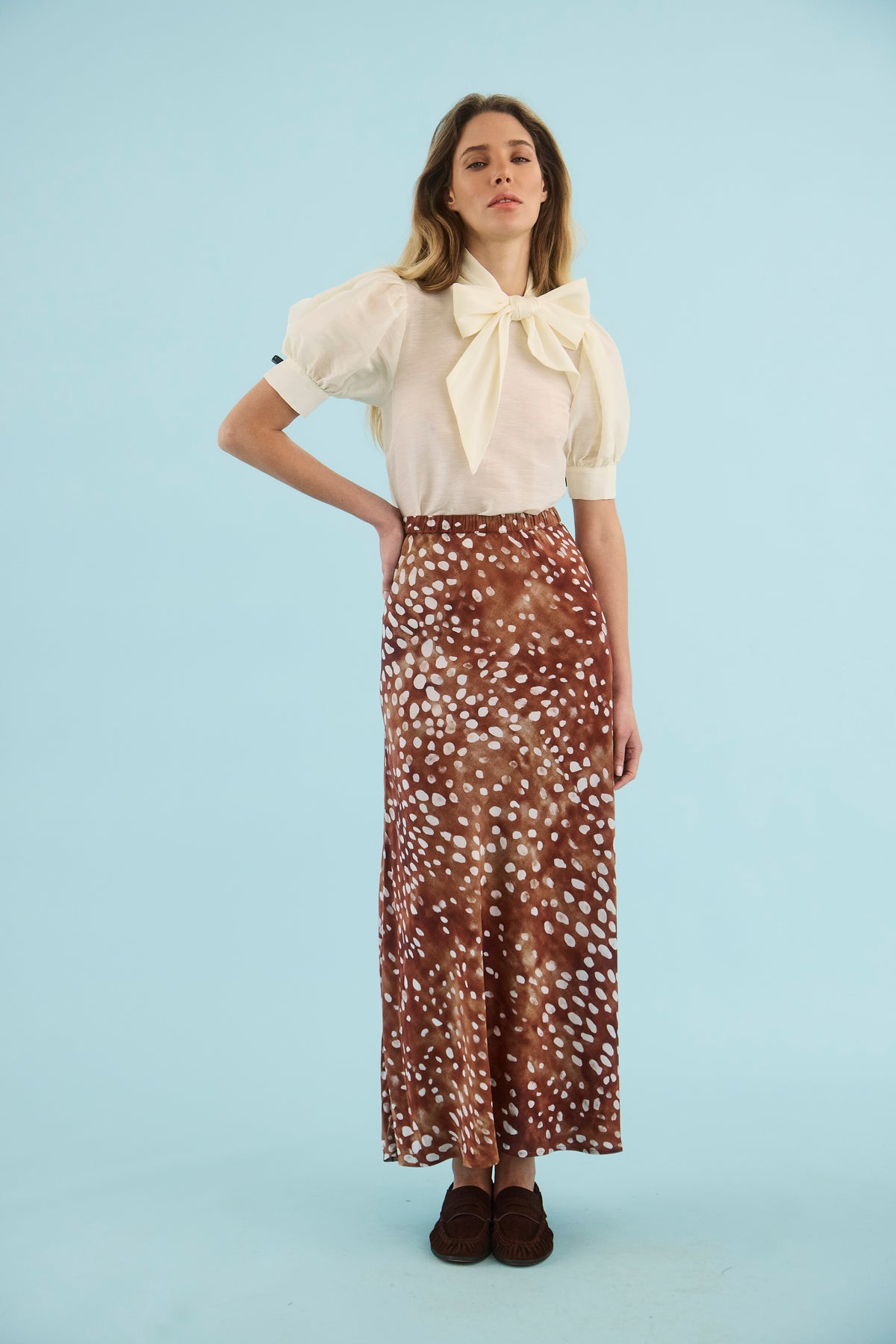 model wearing a dapple doe print skirt with a white top with a bow on a blue background 