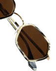 Close-up of a pair of sunglasses with a gold-tone metal frame, dark brown tinted lenses, and a patterned temple arm on a white background.
