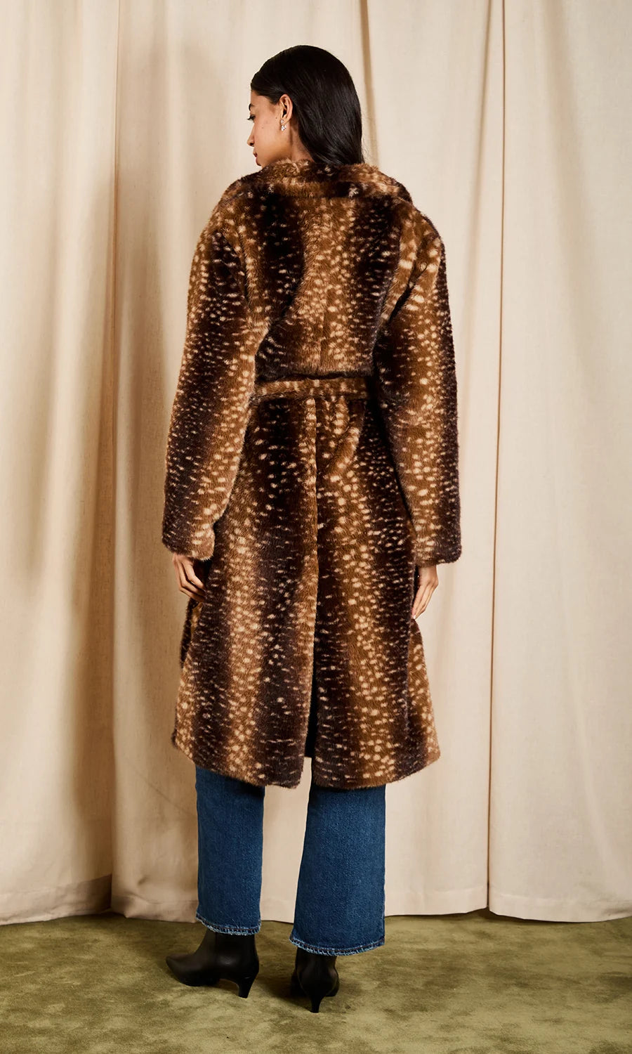 A woman stands with her back to the camera wearing a long brown faux fur coat with a white spotted pattern, blue jeans, and black ankle boots, set against a cream curtain backdrop.