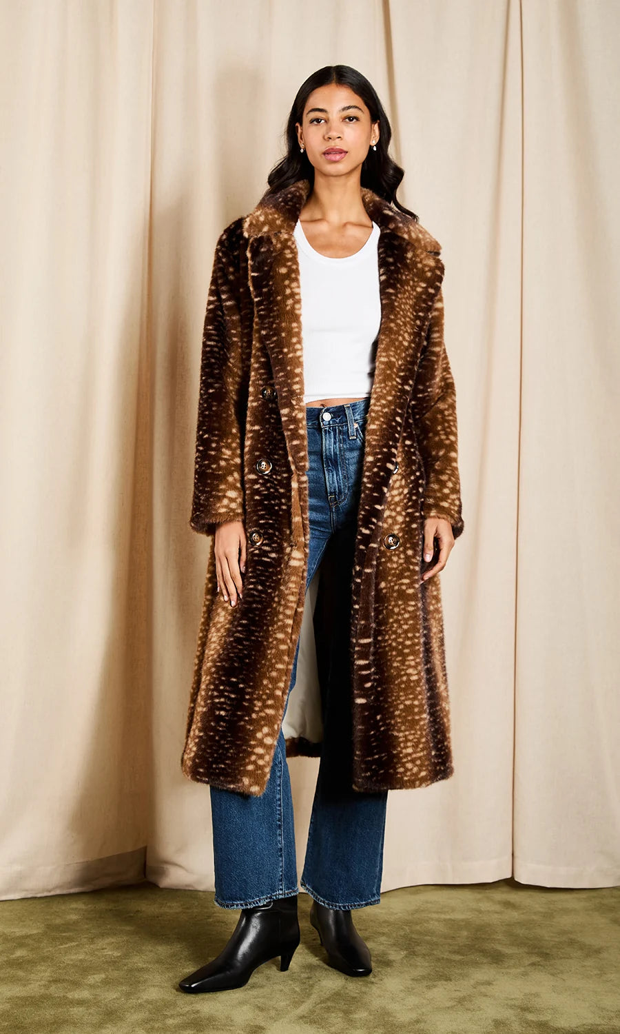 A woman stands in front of beige curtains wearing a long brown faux-fur coat with white speckled spots, a white ribbed top, blue jeans, and black ankle boots.