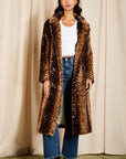 A woman stands in front of beige curtains wearing a long brown faux-fur coat with white speckled spots, a white ribbed top, blue jeans, and black ankle boots.