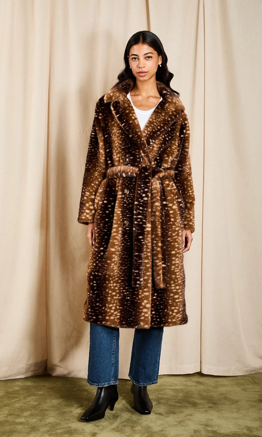 A woman stands in front of a beige curtain backdrop wearing a long brown faux-fur coat with a white speckled pattern, blue jeans, and black ankle boots.