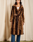 A woman stands in front of a beige curtain backdrop wearing a long brown faux-fur coat with a white speckled pattern, blue jeans, and black ankle boots.
