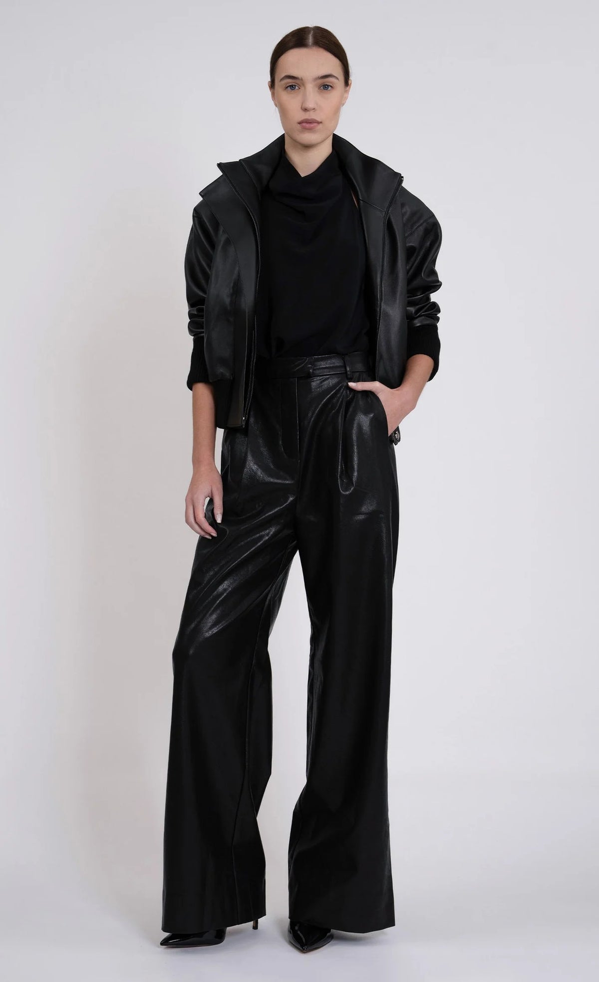 A model stands against a light gray studio background wearing a black faux-leather jacket and matching high-waisted, wide-leg pants with a glossy finish.