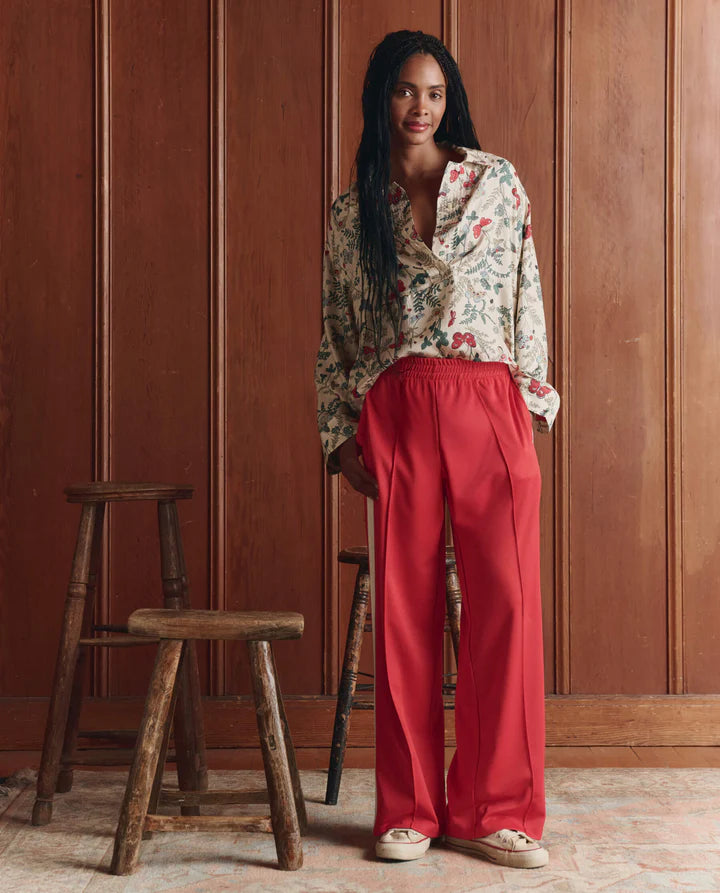 Woman wearing a floral blouse and red pants standing against a wooden paneled wall.