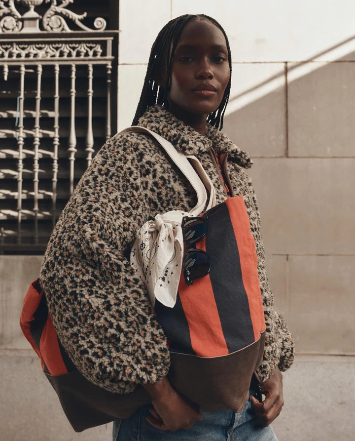 A model wearing a leopard-print teddy coat and blue jeans carries a large Gucci tote bag with orange, navy, and brown stripes, a white bandana, and sunglasses. The background shows a stone wall and a black wrought-iron gate.