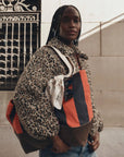 A model wearing a leopard-print teddy coat and blue jeans carries a large Gucci tote bag with orange, navy, and brown stripes, a white bandana, and sunglasses. The background shows a stone wall and a black wrought-iron gate.