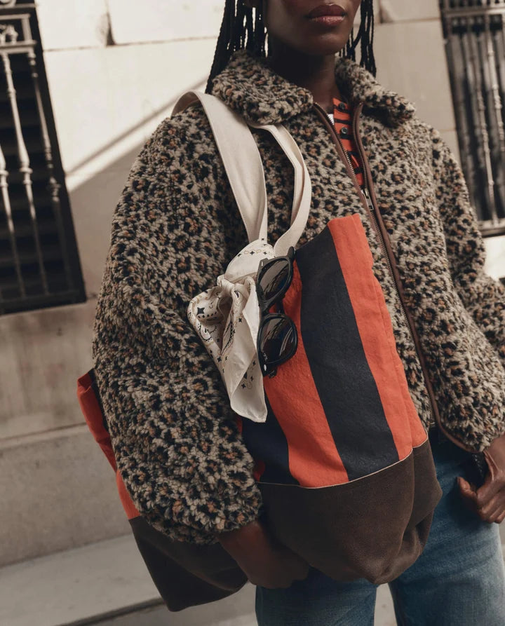 A person wearing a leopard-print teddy jacket and blue jeans carries a large color-blocked shoulder bag with orange, black, and brown panels, a white strap, and sunglasses hanging from the side.