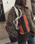 A person wearing a leopard-print teddy jacket and blue jeans carries a large color-blocked shoulder bag with orange, black, and brown panels, a white strap, and sunglasses hanging from the side.