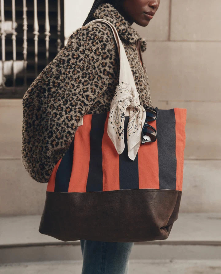 A person wearing a leopard-print coat carries a large striped tote bag with orange, navy, and brown vertical stripes, along with a cream paisley scarf and sunglasses.
