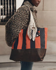 A person wearing a leopard-print coat carries a large striped tote bag with orange, navy, and brown vertical stripes, along with a cream paisley scarf and sunglasses.