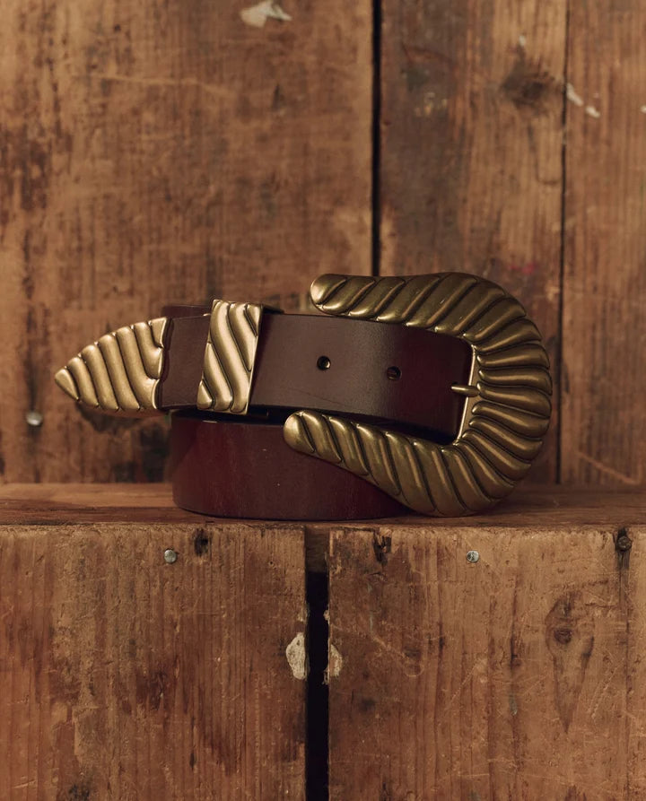 A dark brown leather belt with a large, ornate gold-toned buckle, resting on a rustic wooden surface against a matching wooden plank background.