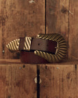 A dark brown leather belt with a large, ornate gold-toned buckle, resting on a rustic wooden surface against a matching wooden plank background.
