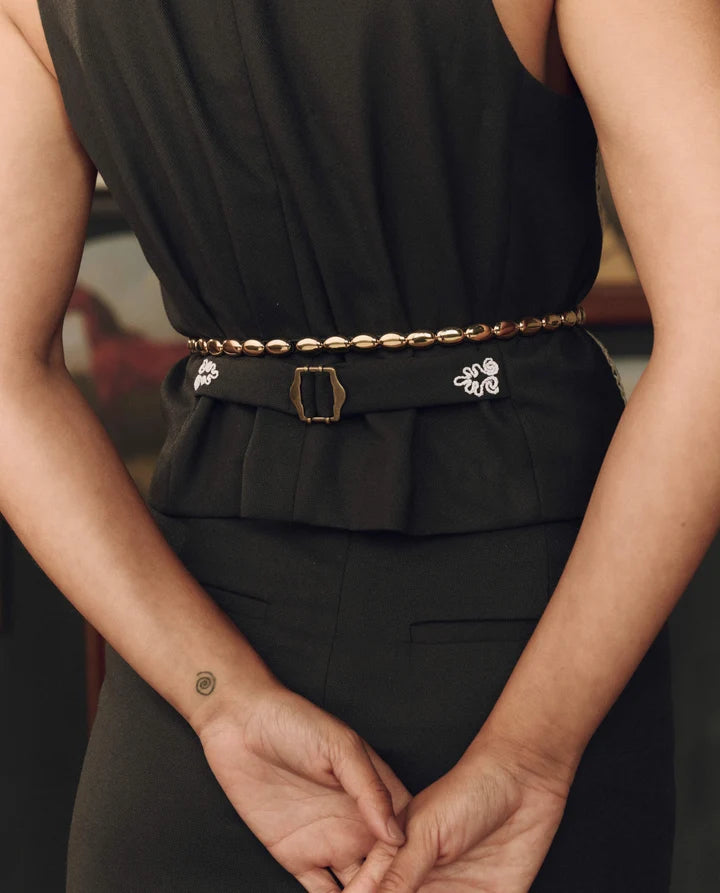 A close-up of a model wearing a black peplum top and black trousers, featuring a gold chain belt with a gold buckle and silver floral embellishments on the sides.