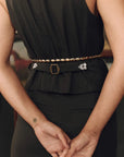 A close-up of a model wearing a black peplum top and black trousers, featuring a gold chain belt with a gold buckle and silver floral embellishments on the sides.