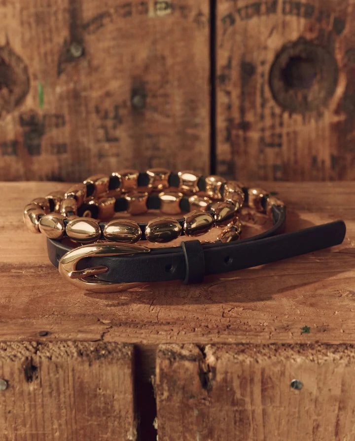 A black leather belt with a large gold buckle and a matching gold chain bracelet, styled on a rustic wooden surface with a matching wooden background.