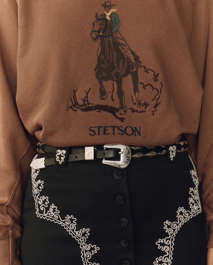 A close-up of a person wearing a brown crewneck sweatshirt with a cowboy riding a horse graphic and the Stetson logo, paired with black western pants featuring white embroidery and a western belt.
