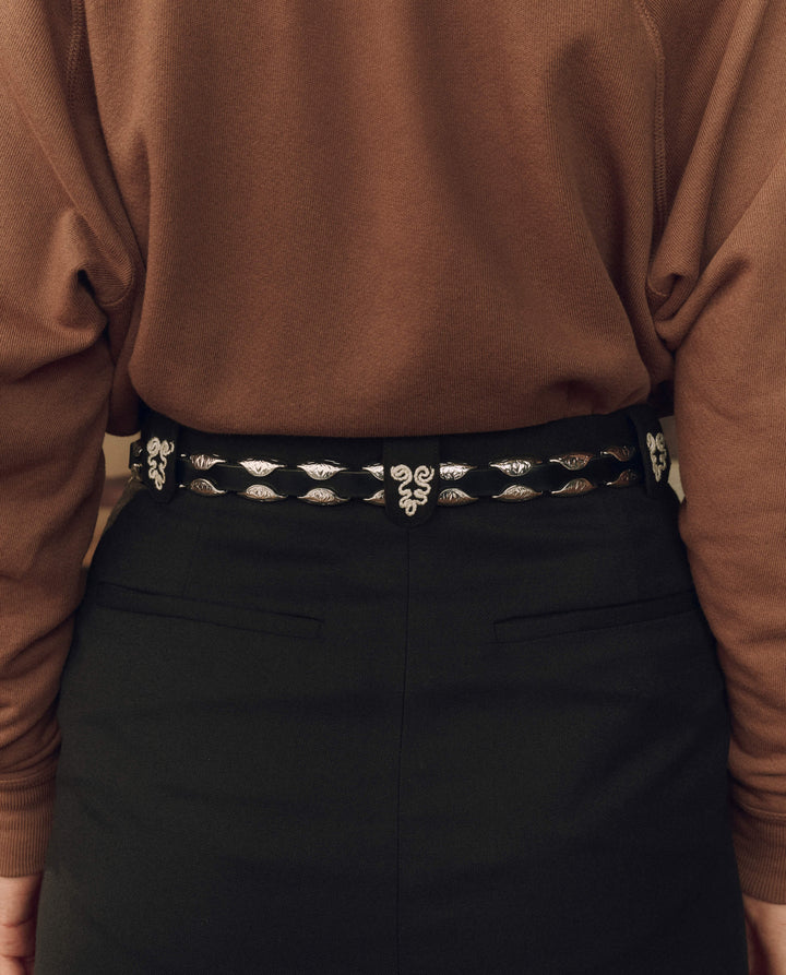 A close-up of a person wearing a black belt with ornate silver-toned woven details and a central decorative buckle, paired with a brown top and dark skirt or trousers.