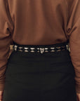 A close-up of a person wearing a black belt with ornate silver-toned woven details and a central decorative buckle, paired with a brown top and dark skirt or trousers.