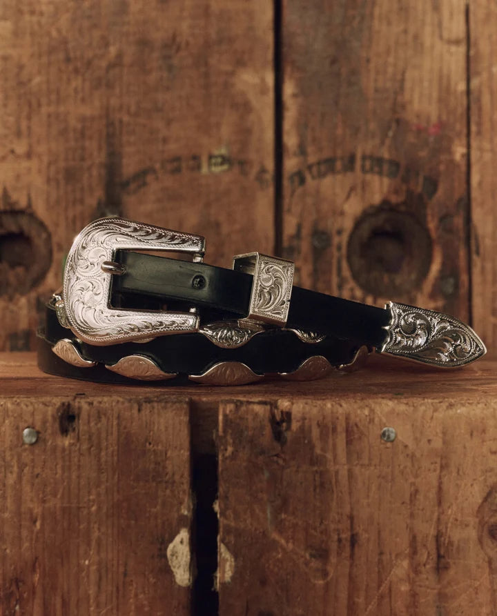 A western-style black leather belt with ornate silver-toned metal hardware, including a large engraved buckle, keeper, and tip, displayed on a rustic wooden surface.