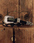 A western-style black leather belt with ornate silver-toned metal hardware, including a large engraved buckle, keeper, and tip, displayed on a rustic wooden surface.