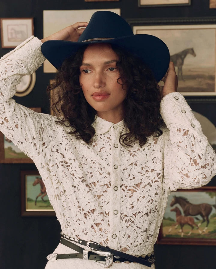 A woman wearing a white lace long-sleeve button-up shirt and a black wide-brim cowboy hat, posing in front of a dark wall with framed horse paintings.