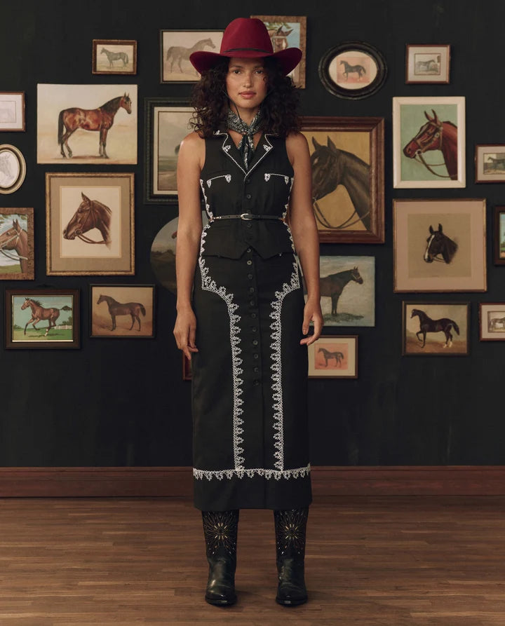 A woman stands in a room with a dark wall covered in framed horse illustrations, wearing a black long-sleeve western-style dress with white embroidered trim, a red wide-brim hat, and black boots.