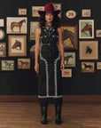 A woman stands in a room with a dark wall covered in framed horse illustrations, wearing a black long-sleeve western-style dress with white embroidered trim, a red wide-brim hat, and black boots.