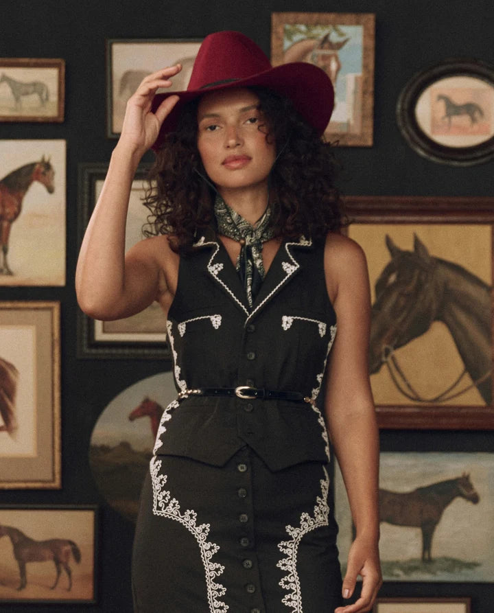 A woman wearing a black western-style sleeveless dress with white embroidered detailing, a matching black belt, and a dark red cowboy hat, standing in front of a wall decorated with framed horse paintings.