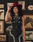A woman wearing a black western-style sleeveless dress with white embroidered detailing, a matching black belt, and a dark red cowboy hat, standing in front of a wall decorated with framed horse paintings.