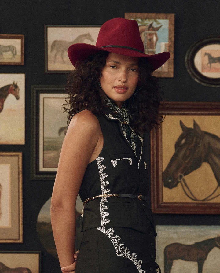 A woman wearing a dark red cowboy hat and a black sleeveless dress with white embroidered details, standing in front of a dark wall decorated with framed horse paintings.