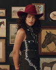A woman wearing a dark red cowboy hat and a black sleeveless dress with white embroidered details, standing in front of a dark wall decorated with framed horse paintings.