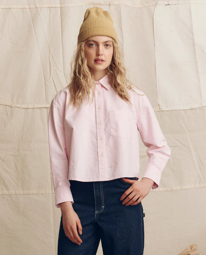 Person wearing a pink shirt, blue jeans, and a beige beanie against a textured beige background
