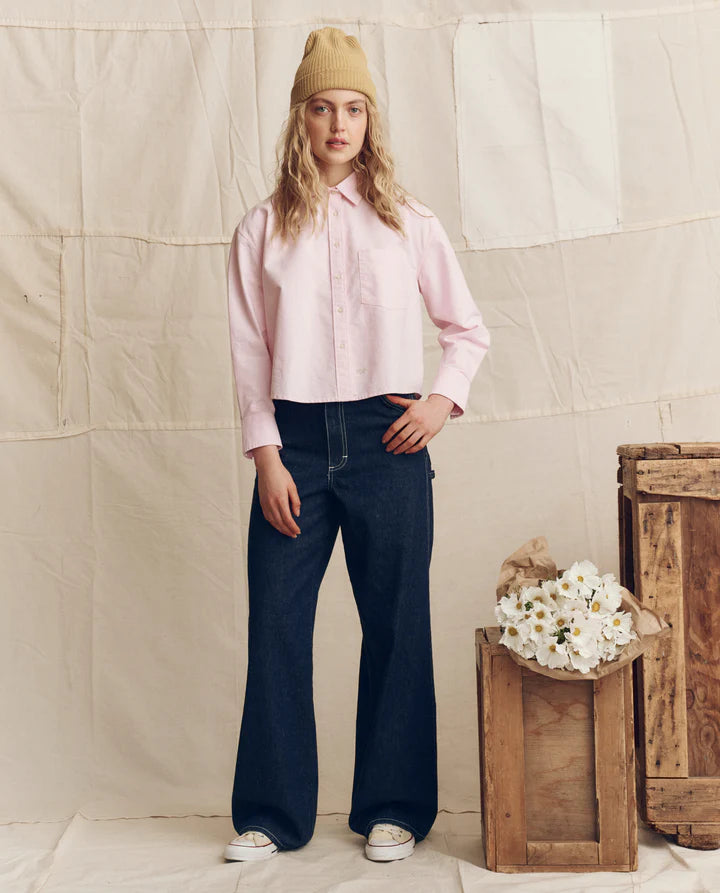 Woman wearing a pink shirt and blue jeans standing next to wooden crates with flowers against a textured wall.