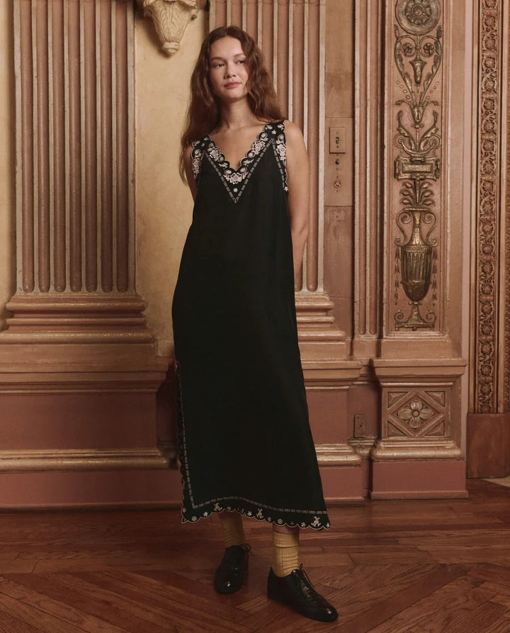 A woman stands in an ornate interior wearing a long black sleeveless dress with embroidered detailing around the neckline and hem, paired with yellow socks and black shoes.