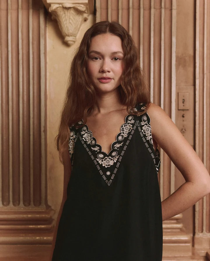 A woman with long wavy brown hair wearing a black sleeveless slip dress with intricate white floral embroidery along the V-neckline, posed in front of a warm-toned classical column background.
