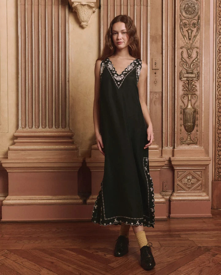 A woman stands in an ornate interior with carved wood paneling and a parquet floor, wearing a sleeveless black midi dress with embroidered detailing around the neckline and hem, paired with beige socks and black shoes.