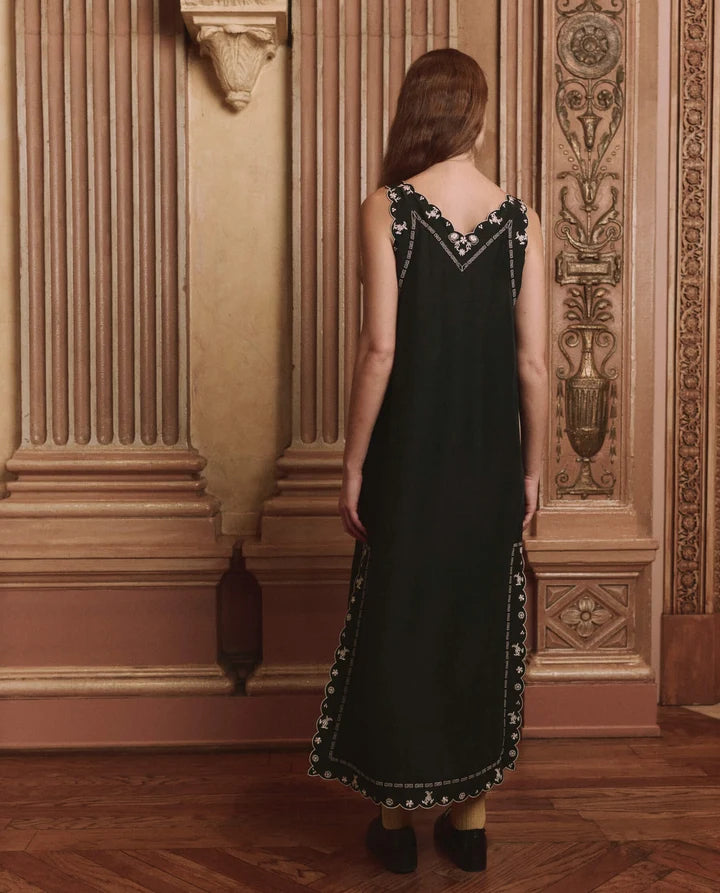A woman stands with her back to the camera wearing a sleeveless black maxi dress with white embroidered detailing along the neckline, armholes, and hem, set against an ornate, warm-toned classical interior wall and wooden floor.