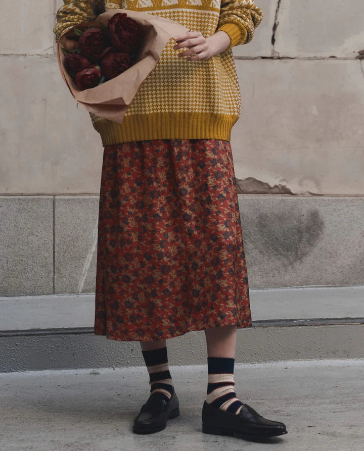 A person standing against a stone wall wearing a mustard yellow knit sweater and a rust-red floral midi skirt, holding a bouquet of dark red roses. Black loafers and striped socks are visible.