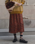 A person standing against a stone wall wearing a mustard yellow knit sweater and a rust-red floral midi skirt, holding a bouquet of dark red roses. Black loafers and striped socks are visible.
