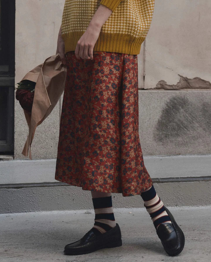 A person wearing a mustard yellow knit sweater and a rust-red midi skirt with a dark floral pattern, paired with striped socks and black loafers, standing outdoors against a textured wall.