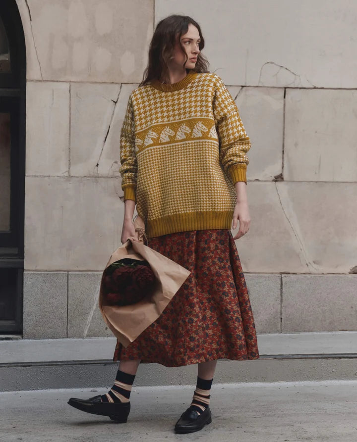 A woman stands outdoors against a stone wall wearing a mustard yellow and white patterned knit sweater, a rust-red floral midi skirt, striped socks, and black loafers, while holding a brown paper bag with a dark floral item inside.