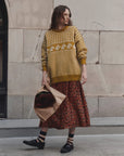 A woman stands outdoors against a stone wall wearing a mustard yellow and white patterned knit sweater, a rust-red floral midi skirt, striped socks, and black loafers, while holding a brown paper bag with a dark floral item inside.
