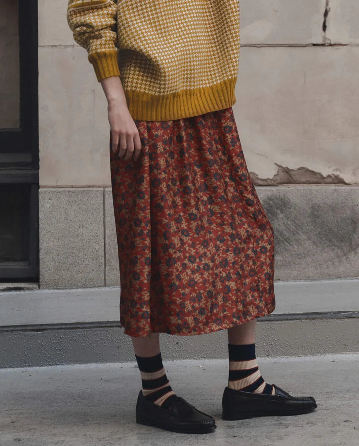 A person wearing a mustard yellow and white checkered knit sweater, a rust-red floral midi skirt, striped socks, and black loafers, standing outdoors against a textured stone wall. side view