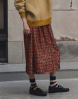 A person wearing a mustard yellow and white checkered knit sweater, a rust-red floral midi skirt, striped socks, and black loafers, standing outdoors against a textured stone wall. side view