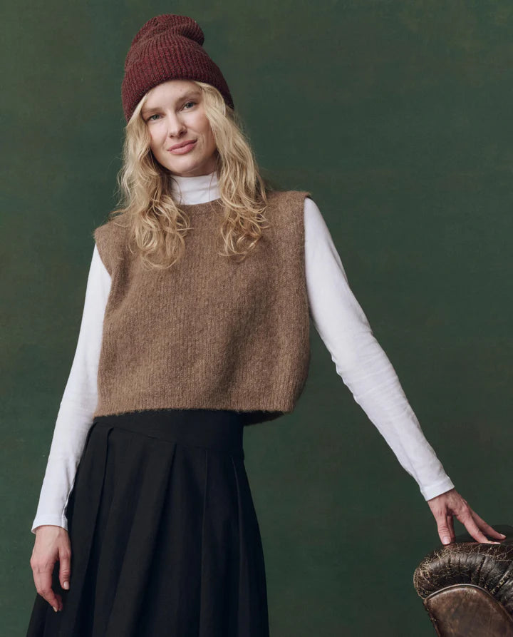 Woman wearing a brown knitted vest, white shirt, and dark skirt against a green background