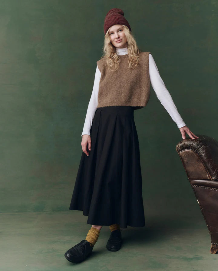 Woman wearing a brown sweater vest, black skirt, and maroon beanie against a green background, full body